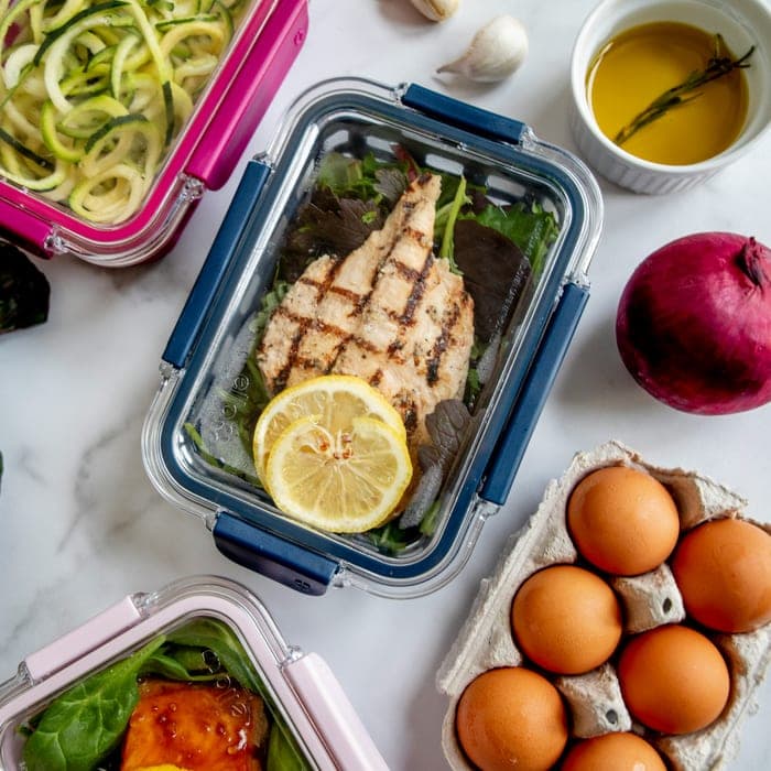 A container of grilled chicken over greens, beside courgette noodles and eggs.