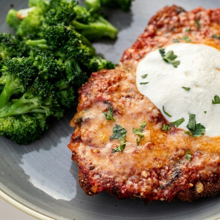 Chicken parmesan topped with melted mozzarella and parsley, with broccoli alongside.