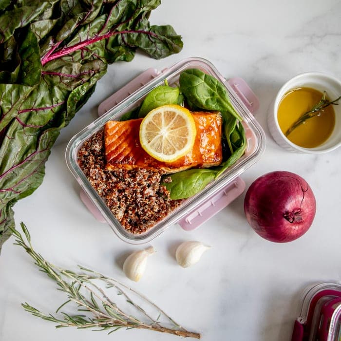 A glass container of glazed salmon with lemon, spinach and quinoa on a marble counter.