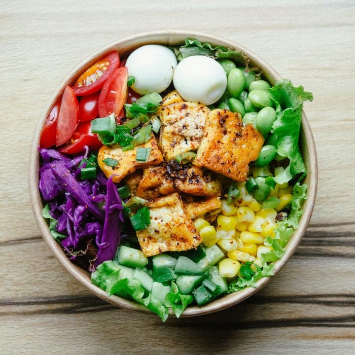 A bowl of crisped tofu with edamame, corn, tomato, cabbage and boiled eggs, seen from above.
